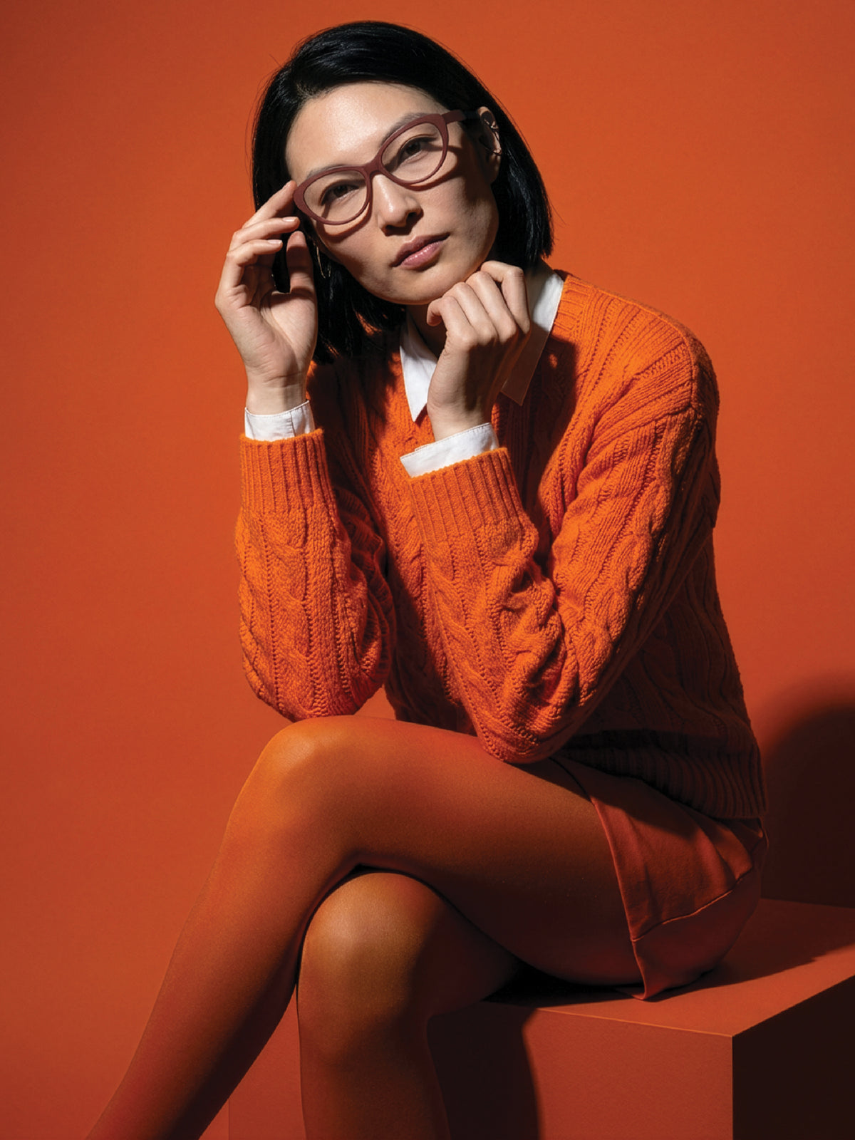 Seated model in an orange cable-knit sweater wearing Blynq Ora rounded glasses in Cordovan