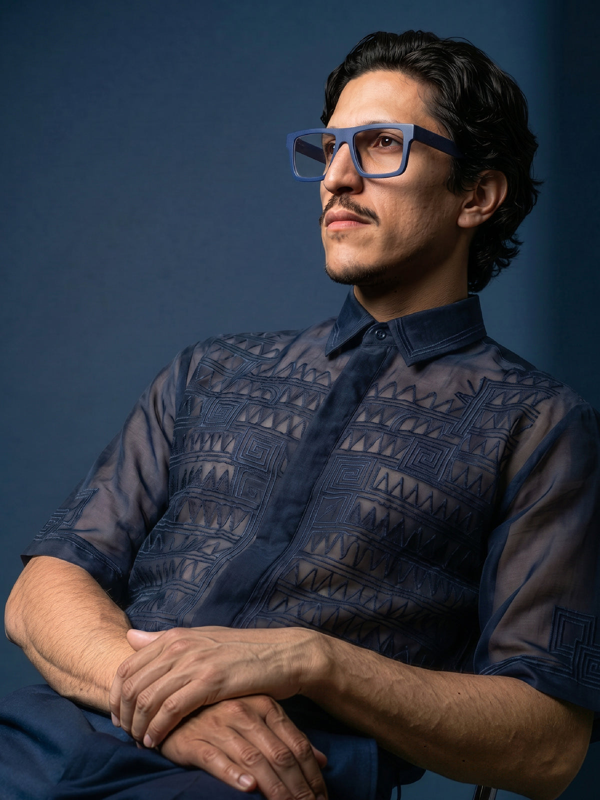 Seated model in a navy sheer shirt wearing Blynq Solo square glasses in Indigo