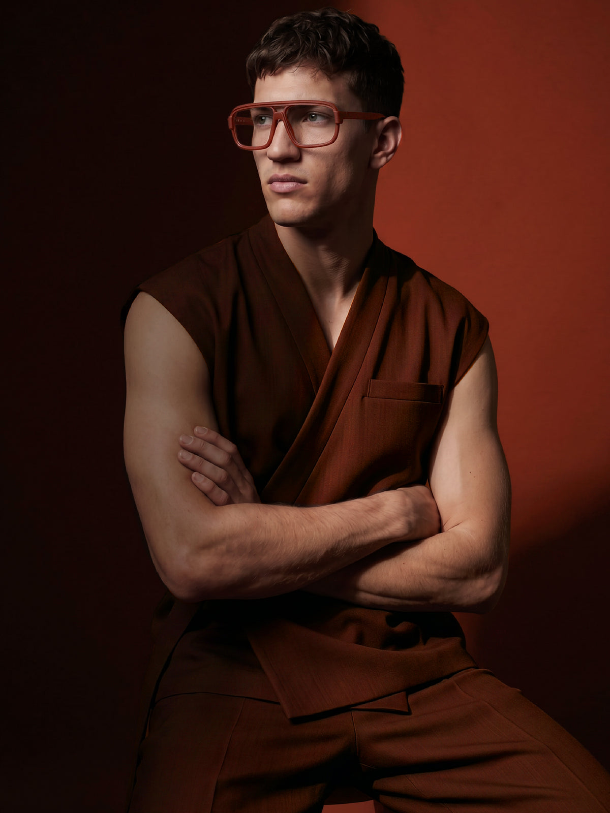 Seated model in a rust sleeveless outfit wearing Blynq Vort double-bridge navigator glasses in Cordovan